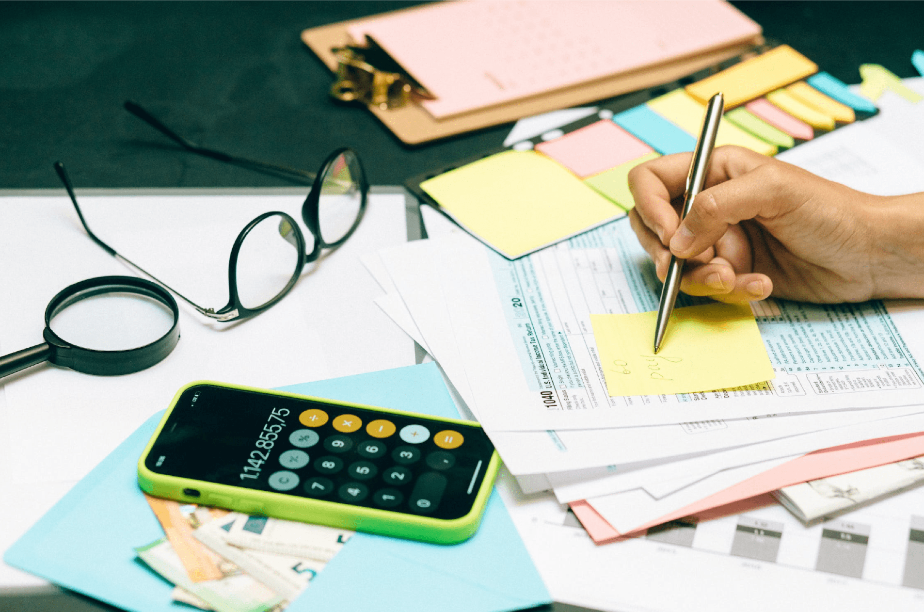 Person signing a stack of papers with an open calculator and glasses laying on the table